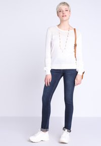 Maglione bianco con dettagli in maglia, abbinato a jeans skinny blu scuro. La modella indossa sneaker bianche e una borsa a tracolla marrone.