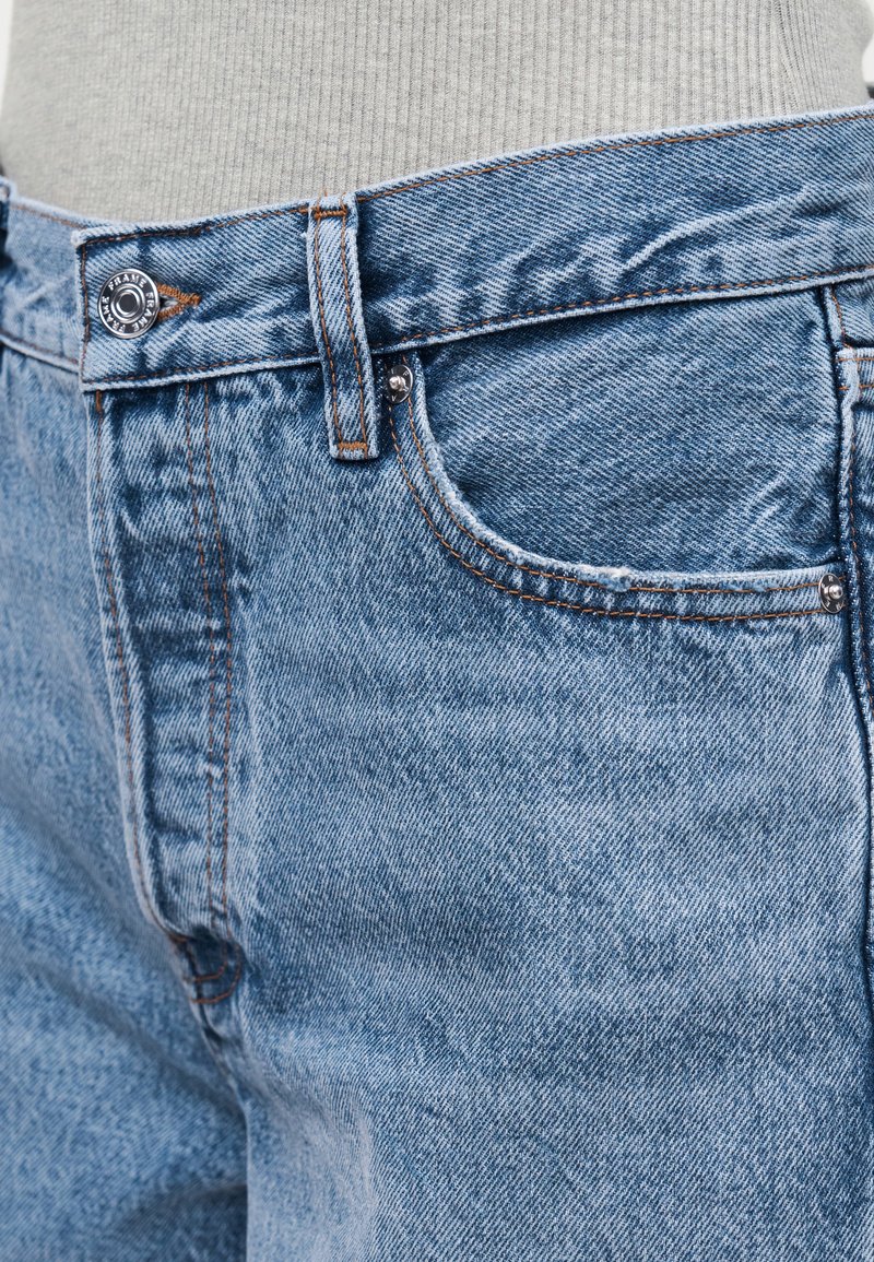 Nahaufnahme der Taille einer blauen Jeans mit Knopf, Gürtelschlaufe und Vordertasche, getragen über einem hellfarbenen, gerippten Oberteil.