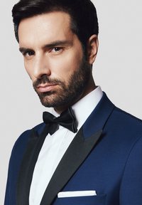 Niebieska marynarka smokingowa z czarnymi satynowymi klapami, biały podkoszulek oraz czarny muszka. Zawiera białą poszetkę. Zbliżenie na twarz modela mężczyzny.