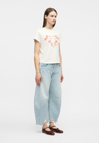 Νεαρή γυναίκα που φοράει λευκό crop top με ροζ γραφικό του Mickey και Minnie Mouse, ανοιχτό μπλε φαρδιά τζιν με σκισίματα και σκούρα καφέ παπούτσια Mary Jane.