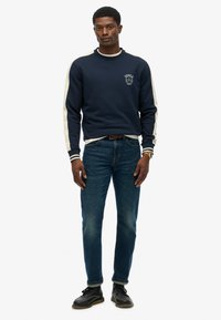Sudadera navy con franjas crema en los hombros y logo en el pecho, combinada con jeans azules y zapatos negros. Ajuste informal y tejido de mezcla de algodón.