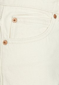 Tecido de denim branco com costura visível e metais em cobre. Possui um bolso frontal com um pequeno bolso para moedas acima dele.