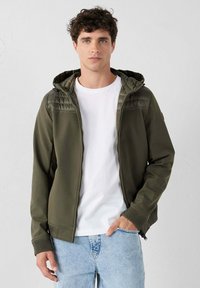 Giacca verde oliva con zip, dotata di cappuccio testurizzato, polsini a coste e due tasche laterali, indossata sopra una maglietta bianca e jeans blu.