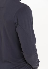 Mörkblå långärmad skjorta med en slät yta, figursydd form och subtil axeldetalj, sedd bakifrån.