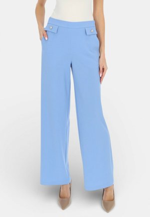 Kobieta ubrana w jasnoniebieskie spodnie z wysokim stanem i szerokimi nogawkami, z patkami na kieszeniach zapinanych na guziki, do tego beżowe szpilki i biały top.