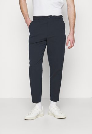 Mann trägt schmal zulaufende marineblaue Hose, weiße Socken, weiße Turnschuhe und ein weißes Kurzarmhemd, steht mit einer Hand in der Tasche.