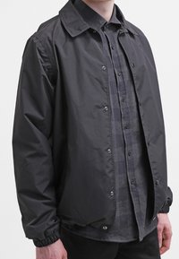 Giacca in nylon nera con colletto, bottoni a scatto, polsini elasticizzati e vestibilità comoda, indossata sopra una camicia a quadri grigia con bottoni.