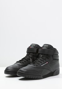 Baskets montantes noires en cuir lisse, avec un design à lacets et une sangle Velcro, dotées d'une semelle texturée et d'accents de marque.