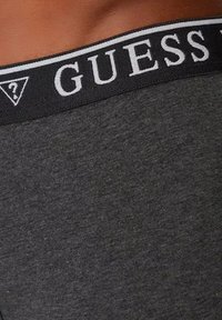 Tissu en coton gris avec une ceinture élastique noire arborant le logo "GUESS" en blanc et un détail graphique en forme de triangle.