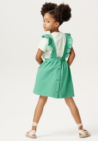 Zielona teksturowana sukienka z szelkami i guzikami z tyłu, w połączeniu z jasną pasiastą koszulą i sandałami na ramiączkach.