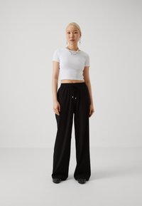 T-shirt blanc côtelé court, pantalon large noir avec cordon de serrage, et chaussures noires avec accents cloutés, le tout sur fond neutre.