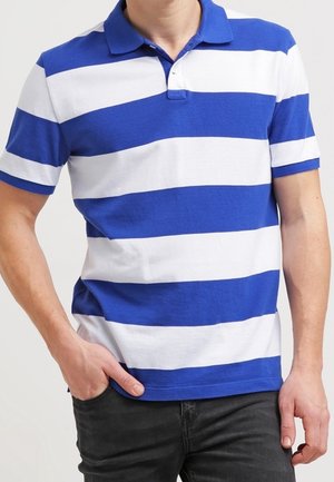 Muž v modro-bílé široce vodorovně pruhované polokošili s krátkým rukávem a límečkem, ruka v kapse, oblečený v černých kalhotách.