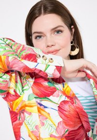 Blazer mit Blumenmuster, der lebendige rote, rosa und gelbe Blumen auf weißem Hintergrund zeigt. Mit einem Reverskragen und goldenen Knopfakzenten.