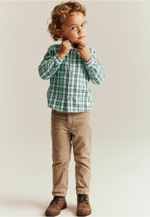 Vihreä ja valkoinen ruudullinen nappipaita, beiget housut ja ruskeat saappaat. Paidassa on kaulus, ja se on valmistettu kevyestä, tekstuurillisesta kankaasta.