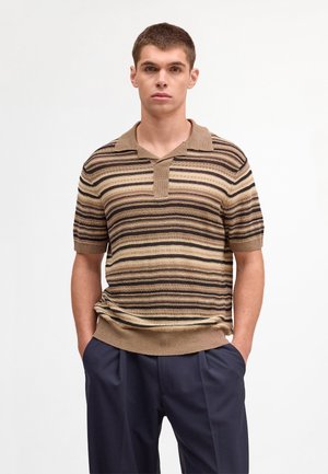 Junger Mann steht mit den Händen in den Taschen, trägt ein braun gestreiftes, kurzärmeliges Strick-Poloshirt und dunkelblaue Hose vor einfachem Hintergrund.