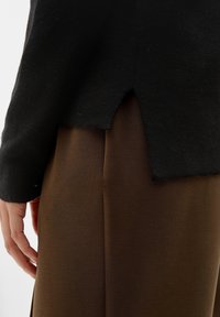 Pull noir avec une finition texturée, doté d'une fente à l'ourlet. Porté avec un pantalon en tissu marron lisse. Design minimaliste, mettant l'accent sur le contraste des couleurs.