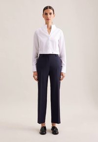 Hvid cropped bluse med ståkrave kombineret med mørkeblå skræddersyede bukser. Modellen bærer sorte loafers. Enkel, ren design.