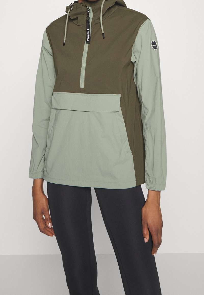 Personne portant une veste à enfiler bicolore verte avec une poche avant et des leggings noirs sur un fond clair uni.