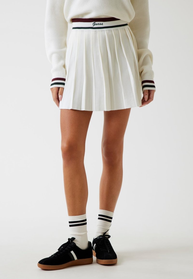 Falda blanca plisada con una cinturilla a rayas en color burdeos y verde que presenta la marca "Guess". La lleva puesta con zapatillas negras y calcetines blancos con dos rayas negras.