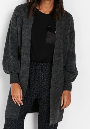 Femme portant un cardigan côtelé gris foncé ouvert devant, un haut noir avec poche et un pantalon noir à cordon avec imprimé léopard, debout devant un fond blanc.