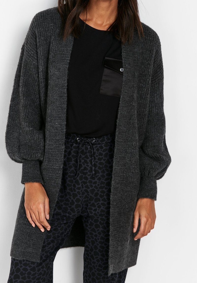 Femme portant un cardigan côtelé gris foncé ouvert devant, un haut noir avec poche et un pantalon noir à cordon avec imprimé léopard, debout devant un fond blanc.