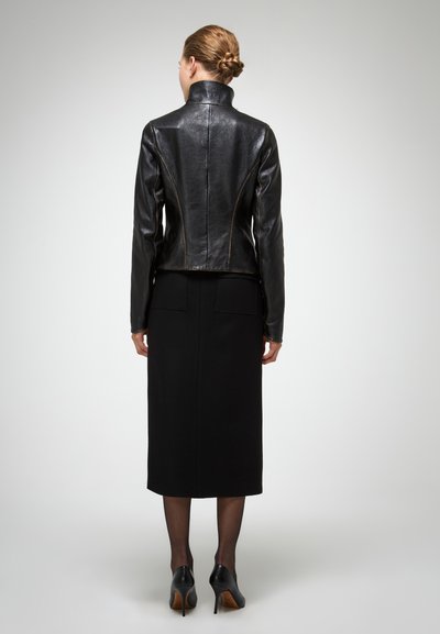 Chaqueta de cuero negra con cuello alto, diseño ajustado y costuras visibles, combinada con una falda negra y zapatos de tacón alto.