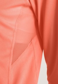 Korallenfarbiges Sportshirt aus glattem Stoff mit atmungsaktiven Netzstoff-Einsätzen an der Seite. Das Design ist eng anliegend und schlank geschnitten.