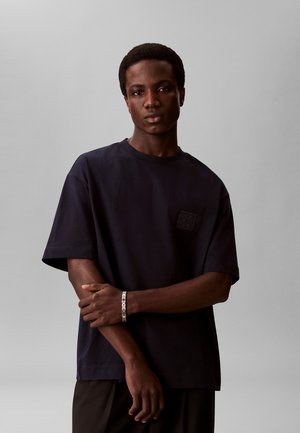 Jonge man met kort haar, die een donker oversized T-shirt en een zilveren armband draagt, staand tegen een effen grijze achtergrond.