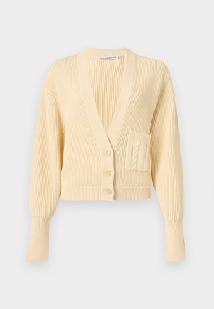 Frau trägt eine kurze blassgelbe Strickjacke mit Knöpfen und hoch taillierte lockere schwarze Hose, steht vor einem schlichten hellen Hintergrund.