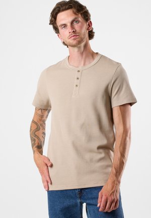 Beige ribbet t-shirt med korte ærmer, rund halsudskæring og tre knapper. Bæret sammen med blå jeans, der viser detaljerede tatoveringer på armen.