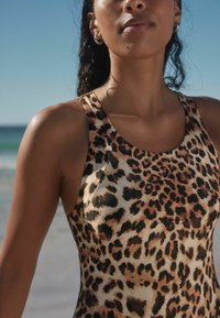 Maillot de bain en imprimé léopard au design ajusté, avec de larges bretelles et une texture lisse, sur fond de plage.