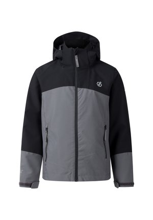 Rothaariger Junge, der eine schwarz-graue Jacke, einen grünen Rucksack, beige Shorts, schwarze Turnschuhe und graue Socken trägt, steht mit neutralem Gesichtsausdruck.
