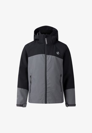 Roodharig jongetje met een zwart-grijze jas, groene rugzak, beige korte broek, zwarte sneakers en grijze sokken, staand met een neutrale uitdrukking.