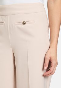 Pantalons beige clair en tissu lisse, dotés de deux poches arrière avec un seul bouton doré. Le design présente des lignes épurées et une coupe ajustée.