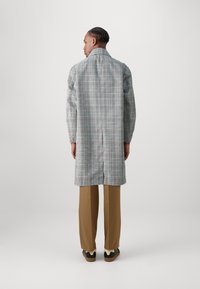 Cappotto lungo a quadri in grigio, blu e bianco, con colletto alto e vestibilità rilassata, abbinato a pantaloni marroni e scarpe nere.
