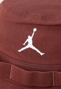 Maroon bucket hat made of fabric; features a white Jordan logo and a textured brim. Includes detailed stitching and a fabric band.