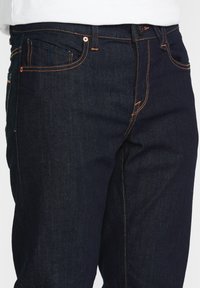 Jeans de mezclilla oscura con costuras naranjas, cinco bolsillos y cierre abotonado. La tela tiene una textura suave y un ligero brillo.