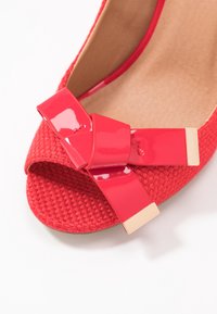 Chaussure ouverte en toile rouge tissée avec un accent de nœud brillant et des détails en or. Texture lisse sur le nœud, contrastant avec le corps tissé.