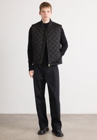 Quilted black vest with a high collar, zip closure, and side pockets, worn over a black long-sleeve top and paired with loose black trousers.