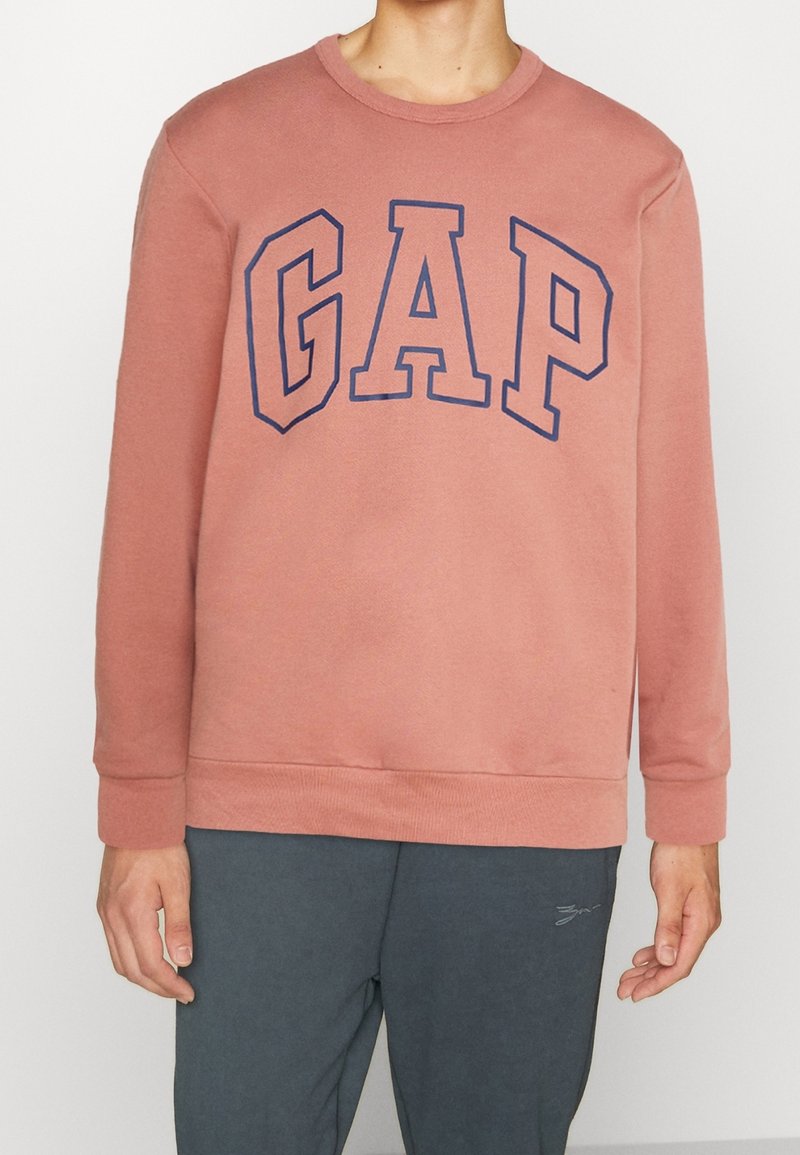 Sudadera rosa de cuello redondo hecha de una mezcla de algodón. Presenta un gran logotipo de "GAP" en color marino en el frontal. Puños y dobladillo ajustados.