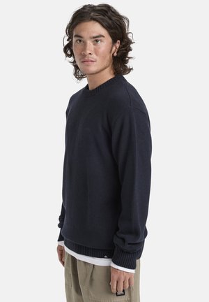 Navy knitted sweater with ribbed cuffs and hem, featuring a small logo on the chest. Layered over a white shirt, worn with beige pants.