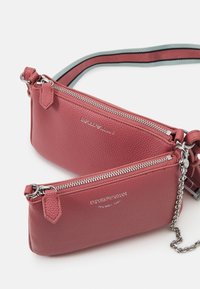 Červená kožená kabelka přes rameno se dvěma kapsami na zip, stříbrnými kovovými doplňky, vyšívaným logem a pruhovaným nastavitelným popruhem.