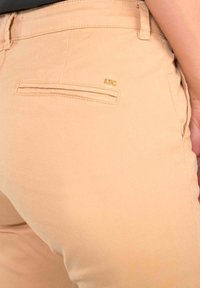 Beige Hose mit einer Gesäßtasche und einem kleinen goldenen "LFC"-Logodetail auf der rechten Seite, getragen von einer Person mit heller Hautfarbe.