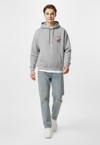 Felpa grigia con tasca frontale, decorata con una grafica a cuore rosso. Abbinata a jeans azzurri chiari e sneakers bianche. Texture liscia.