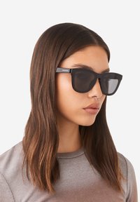 Black rectangular sunglasses with dark lenses, featuring a matte finish and logo detail on the temple. Worn by a model with straight hair.