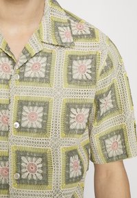 Lyhyt hiha -paita, jossa on vihreä ja beige kukkakroiseedesign, jossa on vaaleanpunaisia ja keltaisia yksityiskohtia. Edessä olevat napit tuovat lisäyksityiskohtia.