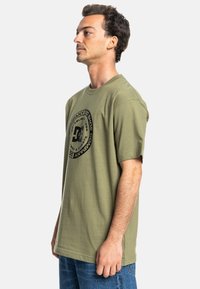 Olivengrünes Baumwoll-T-Shirt mit kurzen Ärmeln, ausgestattet mit einem großen schwarzen runden Logo auf der Vorderseite und einem lockeren Schnitt.