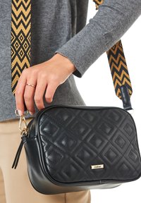Sort quiltet håndtaske med et gyldent logo. Har en tekstureret læderkrop og en multicolorer chevronrem. Vist i hånden.