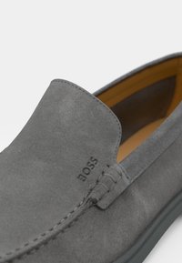 Zapato gris de ante tipo slip-on que presenta un diseño cosido, una marca en relieve y un forro interior suave con suela de goma.