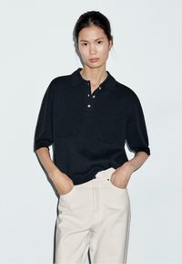 Polo nero a maniche corte con una trama morbida, dotato di pattina con bottoni e due tasche frontali; abbinato a pantaloni beige chiaro.